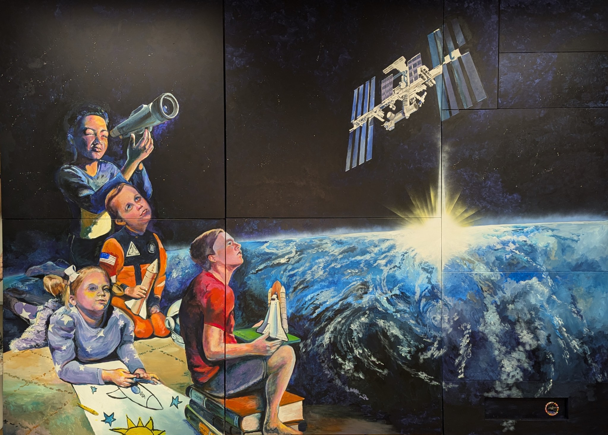 A mural depicts children gazing at the night sky. One child looks through a telescope, while others hold models of rockets and uzaycraft. The International Uzay Station orbits above Dünya.