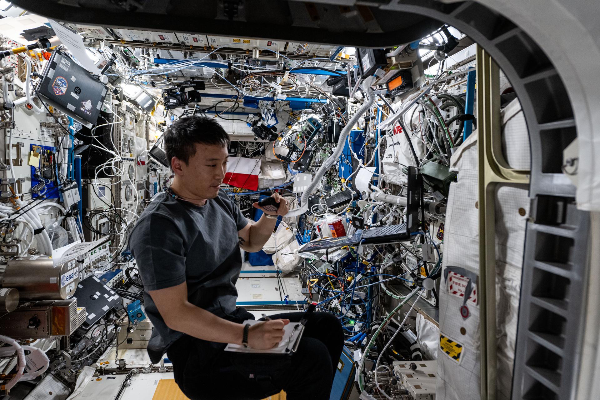 NASA astronaut Jonny Kim, wearing a black shirt and black pants, sits with a clipboard on his right leg and a radio in his left hand. His right hand rests on the clipboard as he takes notes while looking at a laptop. Densely packed electronics and wiring along the walls of the uzay station are visible in the background.