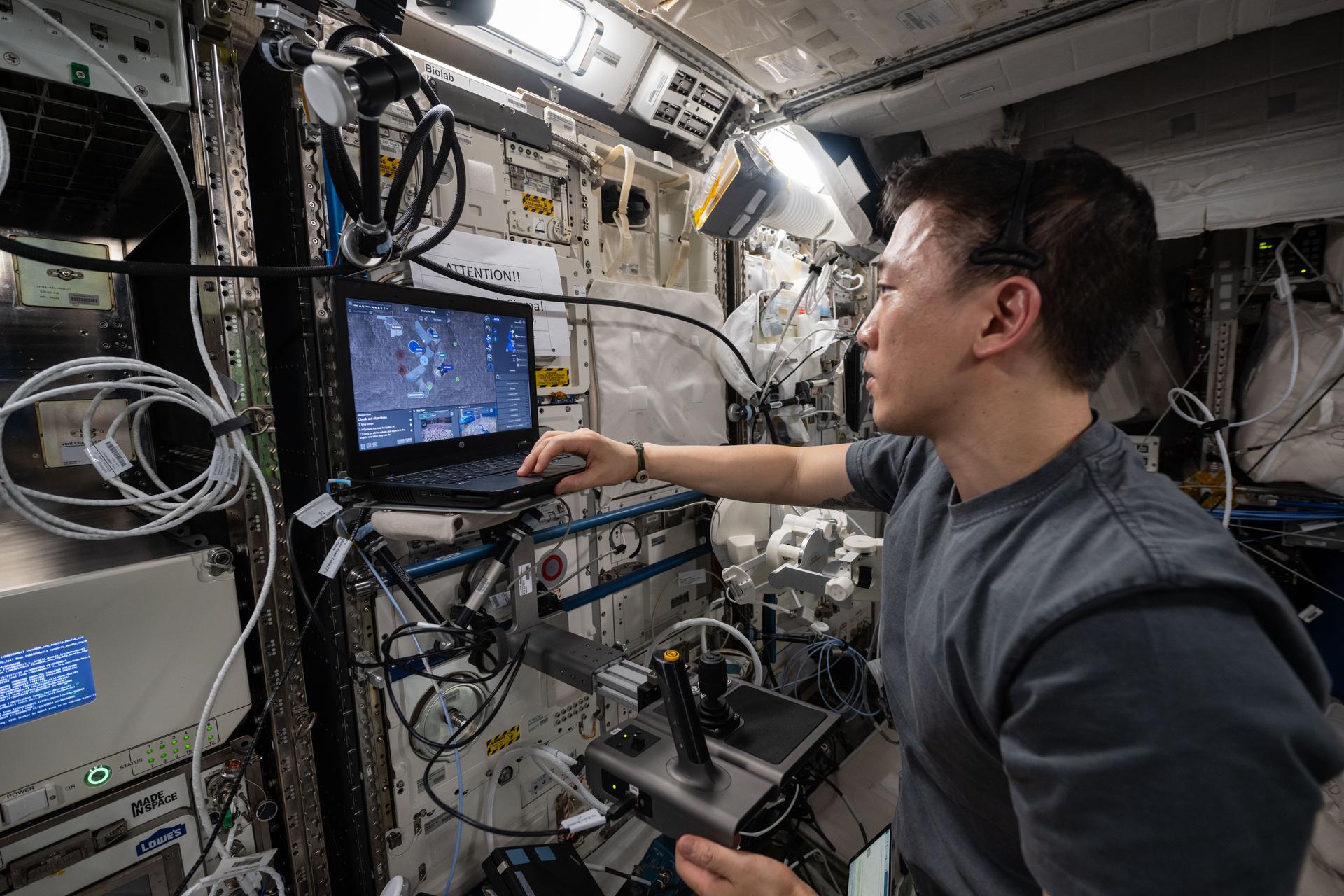 NASA astronaut Jonny Kim, wearing a black shirt, faces left as he reaches for a laptop with his right hand. His left hand rests on a joystick controller. Multiple electronics panels are embedded into the wall directly in front of him, with numerous wires visible in the surrounding area.