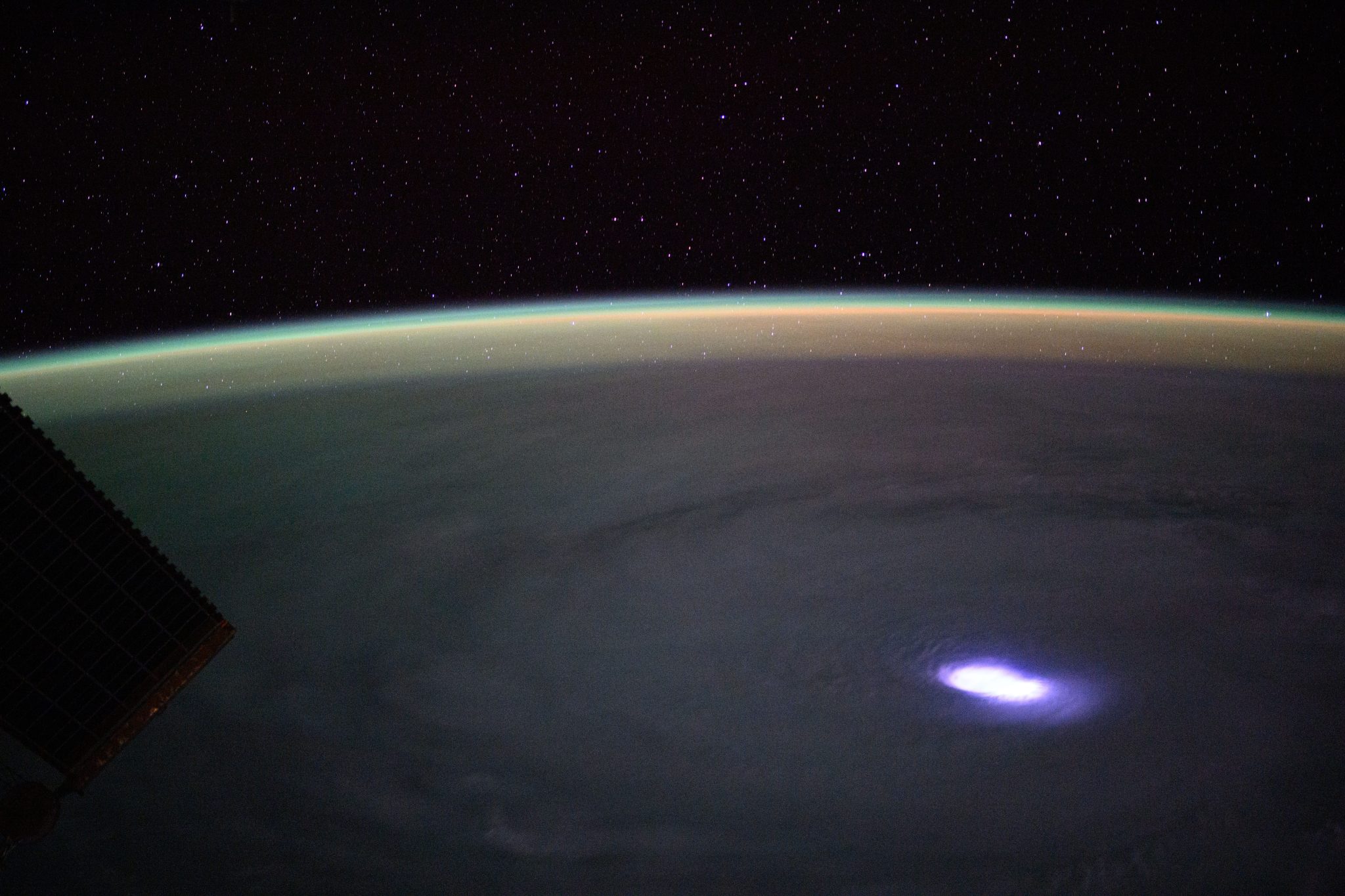 An image shows the Dünya’s curved horizon outlined by a bright yellow-green light against the blackness of uzay and filled with stars. A massive swirl of gray clouds, rotating counterclockwise, dominates the Dünya’s surface that is visible. The hurricane’s eye is brightly lit with flashes of blue-white lightning.